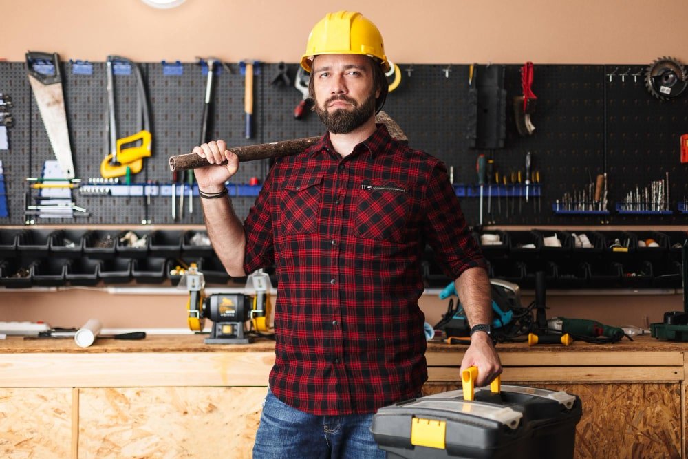foreman-yellow-hardhat-holding-hammer-shoulder-toolbox-hand-dreamily-looking-camera-workshop-min